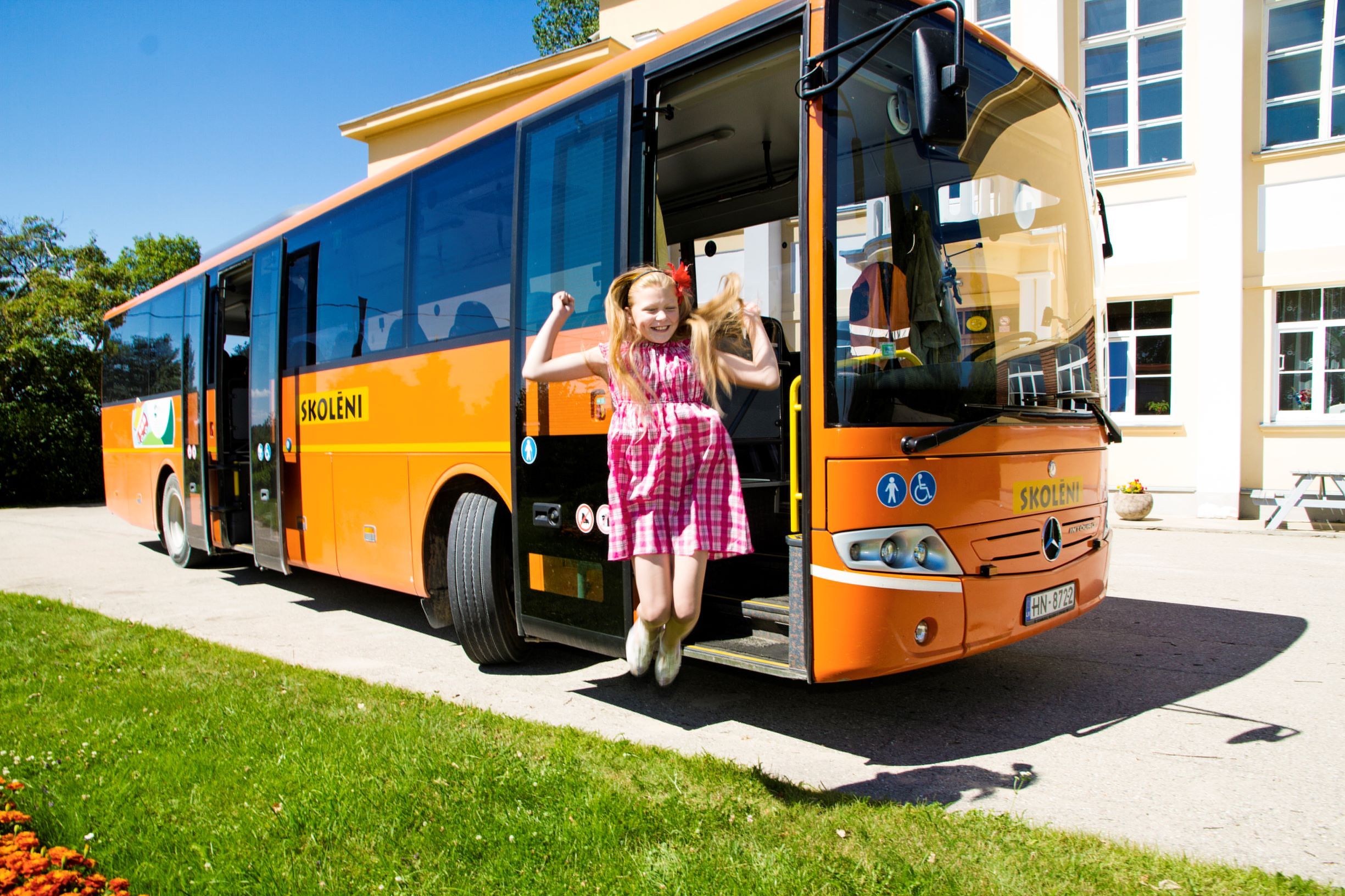Ein fröhliches Mädchen springt aus einem orangefarbenen Schulbus mit der Aufschrift ŠKOLENÍ vor einem hellen Schulgebäude bei Sonnenschein.
