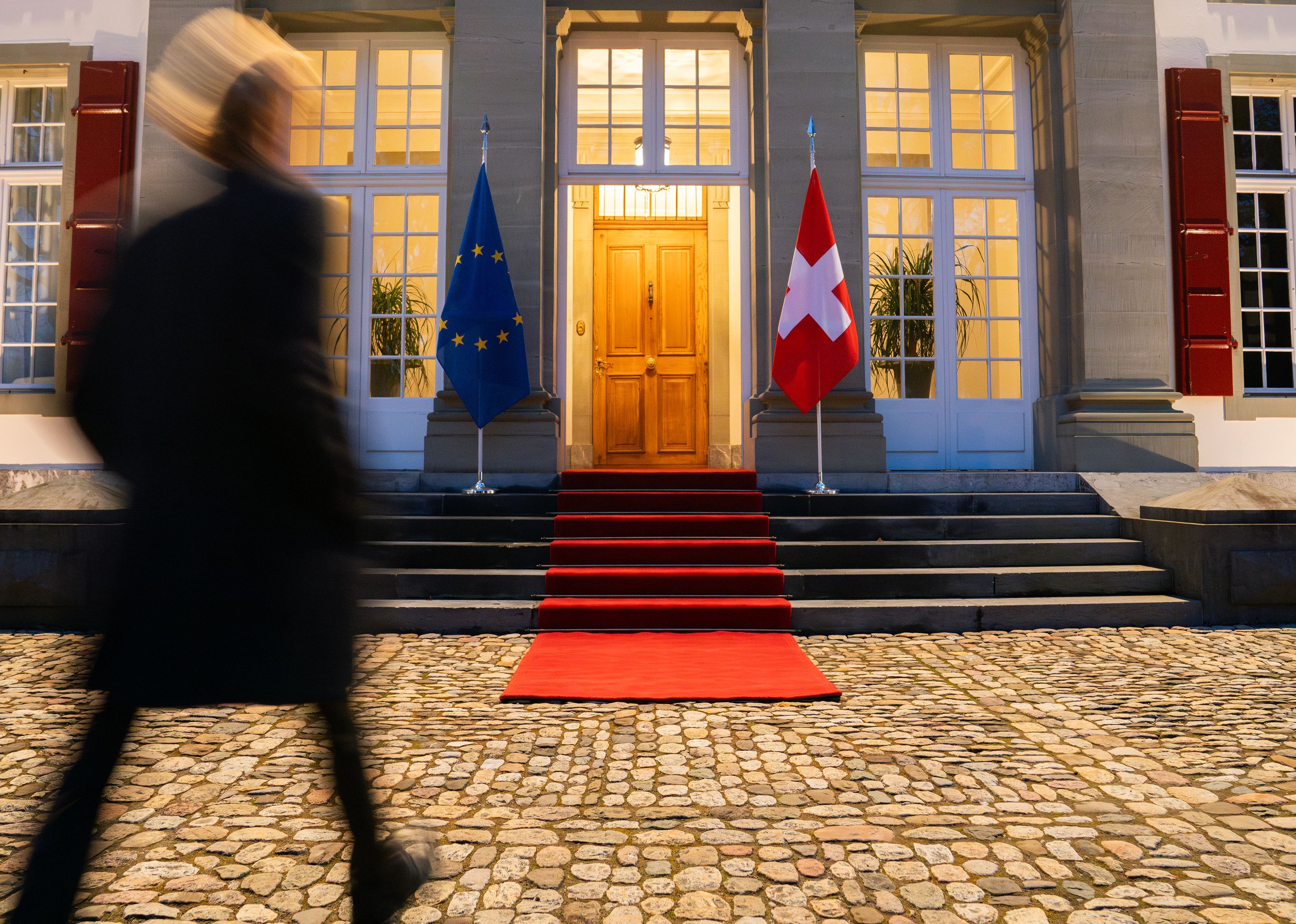 Au Lohn à Kehrsatz, un tapis rouge mène à la porte, à côté de laquelle se trouvent les drapeaux de l'UE et de la Suisse; une personne passe, floue, au premier plan.